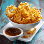 Golden air fryer crispy onions served in a white bowl with dipping sauce on a rustic wood table.